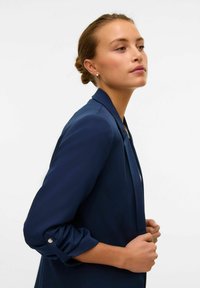Navy blue blazer made of smooth fabric, featuring a tailored design, rolled-up sleeves with silver buttons, and a fitted silhouette.