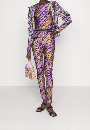 Survêtement violet et doré à motifs avec un haut et un pantalon ajustés, associé à un petit sac rose. Comprend des sandales noires brillantes et élégantes.