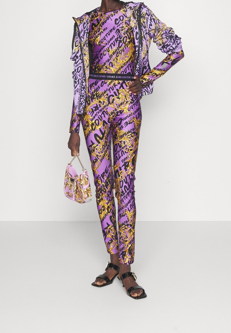 Lila och guld mönstrad träningsdräkt med åtsittande topp och byxor, tillsammans med en liten rosa väska. Utrustad med eleganta, blanka svarta sandaler.