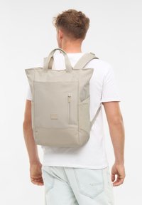 Beige szövet hátizsák téglalap alakú, kettős felső fogantyúval, állítható pántokkal, elülső cipzáras zsebbel és egy kis logóval ellátott folttal.