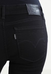 Svarta denimjeans med en slät yta, med en läderpatch i midjan och signaturdetaljer i sömmarna på bakfickan.