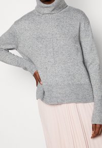 Persoon in een lichtgrijze gemêleerde turtleneck trui met lange mouwen en een lichtroze geplooide rok, hand op de heup.