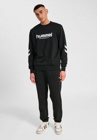 Ung man i svart hummel-tröja och byxor, står med avslappnade händer, vita sneakers med svarta ränder, enkel ljusgrå bakgrund.