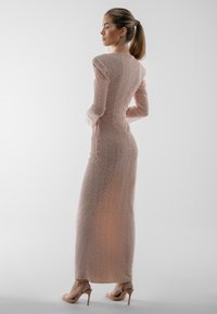 Gara, pieguloša kleita gaiši rozā krāsā ar fakturētu rakstu, garām piedurknēm un siluetu, kas seko ķermeņa līnijām, kombinēta ar brūni bēšiem augstiem papēžiem.