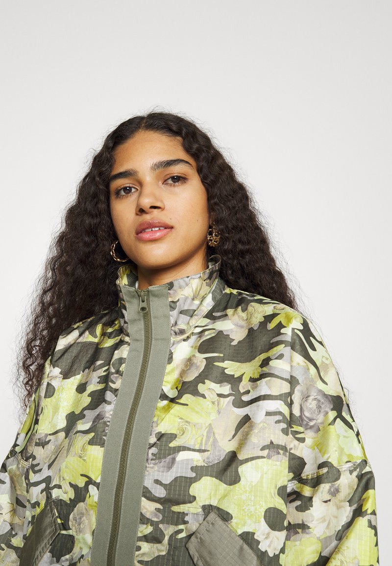 Chaqueta de camuflaje en tonalidades de verde y amarillo, con un alto collar, cierre de cremallera y tejido texturizado. Presenta patrones florales visibles y bolsillos.