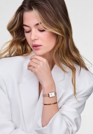 Montre carrée avec un cadran blanc, des garnitures dorées et un bracelet en maille. Accentuée par un fin bracelet doré au poignet. Le modèle porte un blazer blanc.
