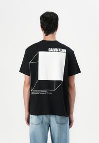 Juodas medvilninis marškinėliai su dideliu baltu geometriniu dizainu ant nugaros ir tekstu "CALVIN KLEIN" viršuje. Klasikinis kirpimas, trumpomis rankovėmis.