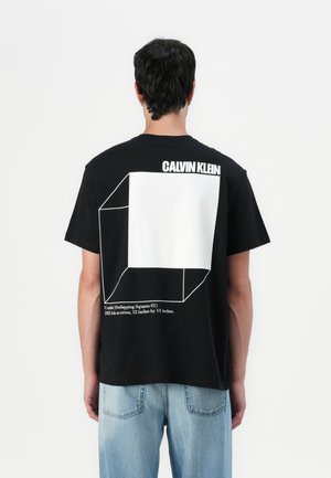 Melns kokvilnas T-kreklis ar lielu baltu ģeometrisku dizainu aizmugurē, virs kura ir teksts "CALVIN KLEIN". Klasisks piegriezums ar īsajām piedurknēm.