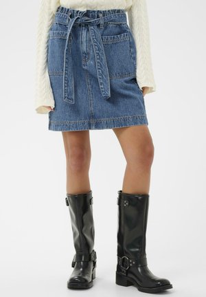 Fustă din denim albastru deschis, cu talie strânsă, două buzunare frontale și tiv cusut. Asortată cu cizme negre din piele până la genunchi.