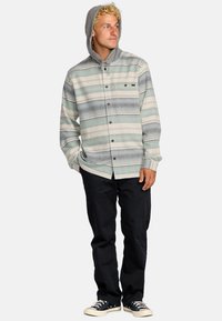 Camisa de franela con capucha en rayas verdes, grises y crema. Presenta botones, un bolsillo en el pecho y una capucha gris en contraste. Se lleva con pantalones negros y zapatillas.