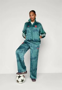 Fato de treino teal com padrão floral, riscas brancas nas mangas e nas calças, acentos em vermelho no logótipo. Em pé sobre uma bola de futebol.