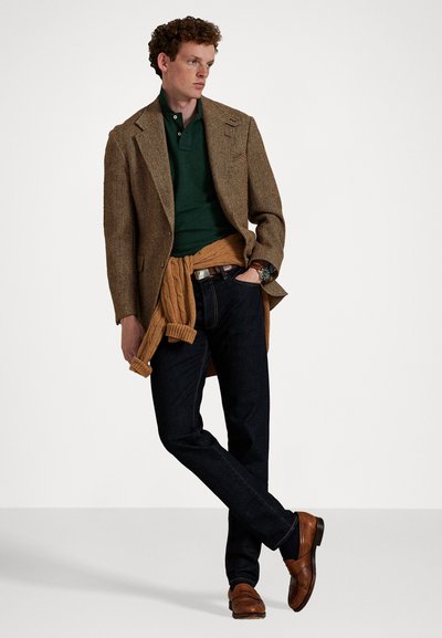 Bruine visgraatblazer over een groene polo, een tan sweater om de taille geknoopt, donkere jeans en bruine loafers. Casual, gelaagde look.