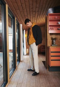 Black bomber jacket, mustard hoodie, cream trousers, and black shoes. Wooden paneling and orange kitchen cabinetry in a modern setting.