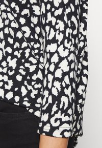 Černobílý top s dlouhým rukávem, který má leopardí vzor, jemně žebrovanou texturu a zaoblený lem. Detail blízko rukávu.