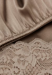 Brun tyg i närbild med en slät, satinliknande textur och tunt nude spetsbård som visar blomstermönster längs kanten.