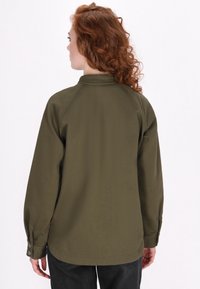 Camicia verde oliva con maniche lunghe e orlo arrotondato. Presenta un colletto alto e polsini con bottoni, mostrando una texture di tessuto liscia e opaca.
