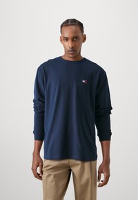 Marineblå langermet T-skjorte, bomullsmateriale, rund hals, liten brodert logo på brystet, avslappet passform, kombinert med beige bukser.