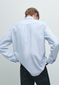 Světle modrá košile s dlouhým rukávem a klasickým límcem, hladká textura a jemný záhyb na zadní straně, zkombinována s tmavými džíny.