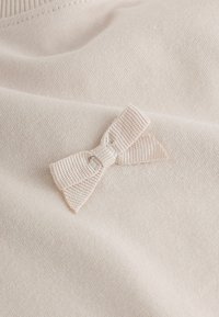 Light beige fabric featuring a small, textured ribbon bow with a striped pattern. The material appears soft and smooth.