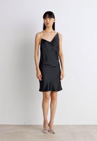 Black satin cowl neck dress with thin straps, knee-length, smooth texture, and a relaxed fit, paired with silver pointed-toe heels.
