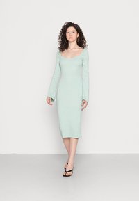 Vestido justo de manga longa em tecido canelado verde menta claro, apresentando decote arredondado e mangas flare. Usado com chinelos pretos.