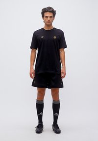 Νέος άνδρας που φοράει μαύρα αθλητικά ρούχα Adidas, συμπεριλαμβανομένου t-shirt, σορτς, καλτσών έως το γόνατο και ποδοσφαιρικών παπουτσιών, μπροστά σε απλό φωτεινό φόντο.