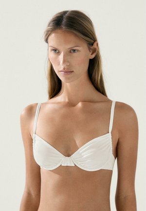 Young woman with long hair wearing a white underwire bikini top against a plain light background.