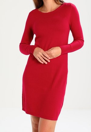 Kobieta w dopasowanej czerwonej sukience sięgającej do kolan, z długimi rękawami zakończonymi prążkowanymi mankietami, stojąca z rękami złożonymi przed sobą na jednolitym tle.