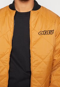 Veste bomber matelassée orange avec un col noir côtelé. Comprend un logo "OBEY" brodé sur la zone gauche de la poitrine, assortie à une chemise noire en dessous.