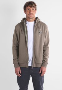 Hoodie zippé marron avec une doublure intérieure texturée, comportant un petit détail graphique sur la poitrine. Il a des cordons de serrage et des poignets côtelés.