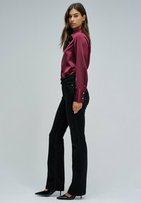 Blusa de satén burdeos con mangas largas y acentos de botones dorados, combinada con jeans de campana negros y tacones negros puntiagudos. Fondo liso claro.