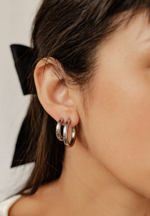 Drei miteinander verbundene silberne Creolenohrringe, die an einem Ohr gezeigt werden, mit einer polierten Oberfläche und einem glatten, abgerundeten Design.