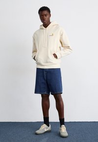 Beige hættejakke med frontlomme og snor, parret med denimshorts. Iført grå og beige sneakers, stående på et blåt tæppe.