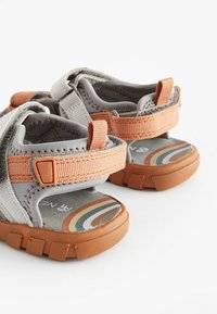 Grå og orange sandaler til børn med en struktureret overflade, justerbare stropper og gummisåler med et farverigt regnbuemønster på indersålen.