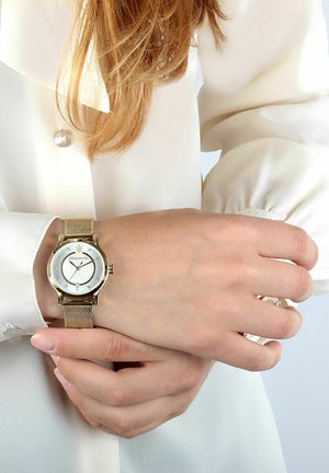 Orologio con cinturino a maglia dorata, con quadrante rotondo bianco, dettagli argentati e indicatori simili a diamanti. Indossato su un polso contro una blusa chiara.