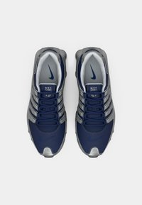 Modré a šedé tenisky Nike Shox s elegantným dizajnom, obsahujúce textúrovaný zvršok, výrazný systém šnurovania a viditeľné akcenty značky.