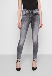 Jeans skinny de mezclilla gris con textura desgastada, cinco bolsillos y cierre de botón. Combinados con tacones negros de punta y una camiseta sin mangas oscura.
