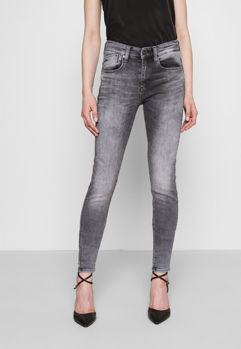 Jeans skinny de mezclilla gris con textura desgastada, cinco bolsillos y cierre de botón. Combinados con tacones negros de punta y una camiseta sin mangas oscura.
