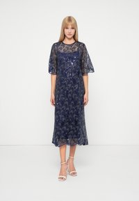 Robe en dentelle navy avec des sequins, manches courtes bouffantes et ourlet festonné. Elle présente un corsage ajusté avec une jupe fluide, créant une silhouette élégante.