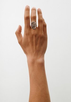 Vasen käsi, jossa on keskivaalea ihon sävy, käyttää hopeista ovaalista sormusta, jossa on musta geometrinen sisustus keskisormessa valkoista taustaa vasten.