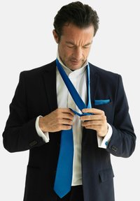 Cravate bleue en tissu texturé, portée par une personne en costume noir avec une chemise blanche, ajustant la cravate autour de son cou.