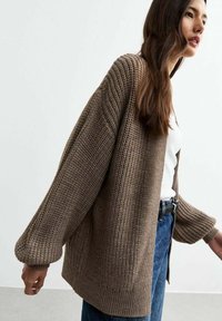 Cardigan lavorato a maglia in marrone terroso, con maniche oversize, vestibilità rilassata e un motivo testurizzato. Indossato sopra una maglietta bianca e jeans blu.