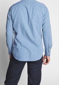 Ljusblå långärmad skjorta med en mjuk textur, knäppning vid ärmarna och en rundad fålldesign. Bakifrån syns sömdetaljer och en skräddarsydd passform.