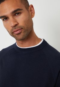 Maglione lavorato a maglia blu navy con colletto a girocollo e una breve fodera bianca interna. Presenta un tessuto texture e un design a maniche raglan per il massimo comfort.