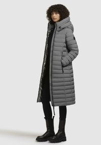 Gray puffer coat with a hood, featuring a quilted design, front zipper, elastic cuffs, and a shiny inner lining. Black chunky boots visible.