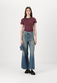 Bordeauxrode t-shirt gecombineerd met lichtblauwe, wijde denim jeans met een gestrikte tailleband. Het model houdt een bijpassende blauwe accessoire vast. Zwarte schoenen maken de look compleet.