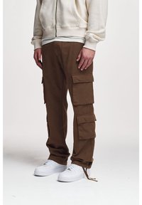 Hnědé pracovní kalhoty s množstvím kapes, volným střihem a manžetami. Kombinovány s bílými teniskami a světlou mikinou s kapucí.
