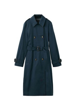 Femme aux longs cheveux bouclés foncés, portant un trench-coat foncé ceinturé, regardant droit devant sur un fond uni.