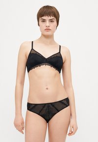 Ensemble de lingerie noire comprenant un bralette transparent avec de la dentelle et des bords festonnés, associé à des culottes assorties à motifs. Design minimaliste.