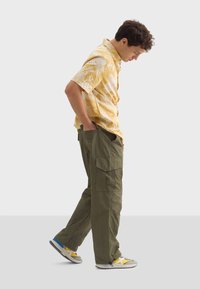 Korte mouwen gele shirt met witte palmbladprint, gecombineerd met olijfgroene cargobroek met zijzakken en een losse pasvorm. Casual sneakers.